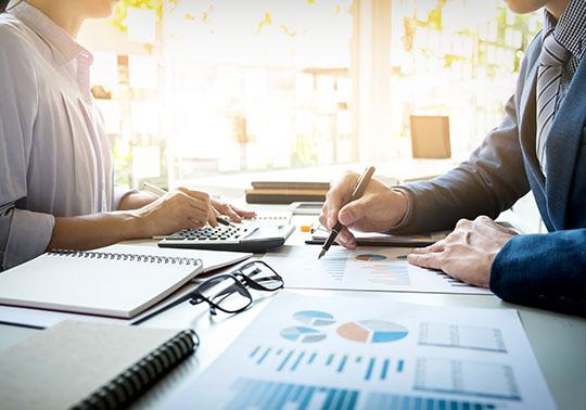 Zwei Mitarbeiter sitzen sich an einem Schreibtisch gegenüber und besprechen die auf dem Tisch liegenden Statistiken und Bilanzen eines Mandanten