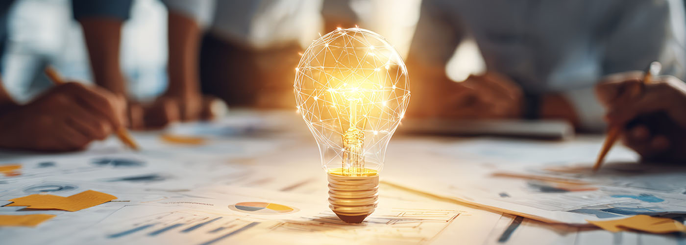 Bildmontage einer Glühbirne  als Zeichen von Ideenentwicklung steht auf einem Tisch mit ausgedruckten Bilanzen und Statistiken. Um den Tisch herum sitzt das Steuerberatungsteam.