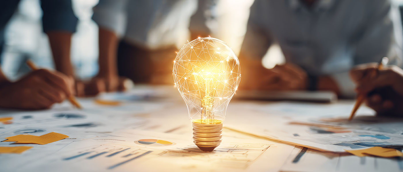 Bildmontage einer Glühbirne  als Zeichen von Ideenentwicklung steht auf einem Tisch mit ausgedruckten Bilanzen und Statistiken. Um den Tisch herum sitzt das Steuerberatungsteam.